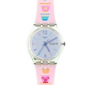 May include: A Swatch watch with a light blue face, featuring the day of the week and the number 18. The watch has a clear, textured bezel and a pink strap adorned with teacup designs. The watch hands are blue and purple.
