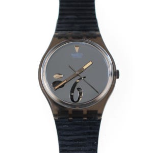 May include: A Swatch watch with a dark gray face and a black strap. The watch face features gold-colored hands and the Swatch logo. The watch has a transparent, dark gray case and a black, ribbed strap.