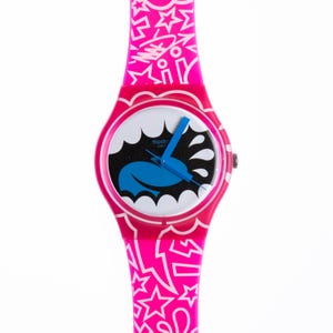 May include: A pink Swatch watch with a white face and a blue and black graphic of a mouth with water splashing out. The watch band has a white and pink pattern with stars and other geometric shapes.