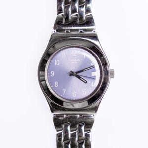 May include: A silver-toned Swatch watch with a light purple face and matching hands. The watch has a linked metal band and a small dial on the side. The watch face displays the Swatch logo and the word "Swiss".