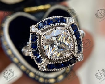 May include: A square-shaped diamond engagement ring with a halo of smaller diamonds and a border of blue sapphires. The ring is set in a silver band.