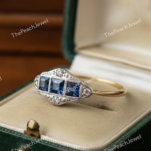 May include: A vintage-style ring with three square, deep blue gemstones set in a silver-colored metal. The ring has a gold band and is displayed in a green velvet jewelry box. Small clear stones accent the blue gems.