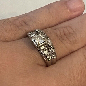 May include: A silver engagement ring and wedding band set. The engagement ring features a square-cut center stone and smaller stones on the band. The wedding band has a matching design with smaller stones. The rings are worn on a person's finger.