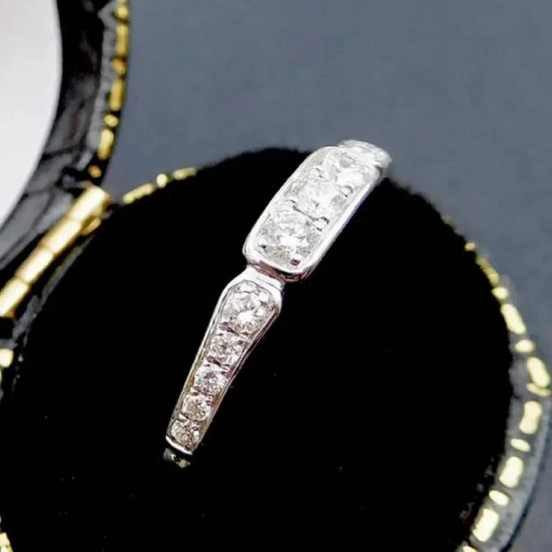 Puede incluir: Un delicado anillo de banda de plata con una fila de peque&ntilde;os diamantes brillantes engastados en un engaste de canal. El anillo se muestra en una caja de anillo de terciopelo negro.