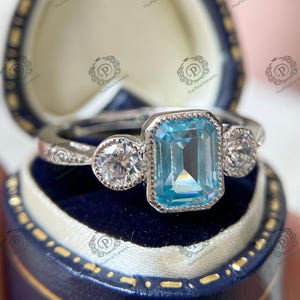 May include: A silver ring with a large, rectangular, light blue gemstone in the center, flanked by two smaller, round, clear gemstones. The ring is set in a blue jewelry box.