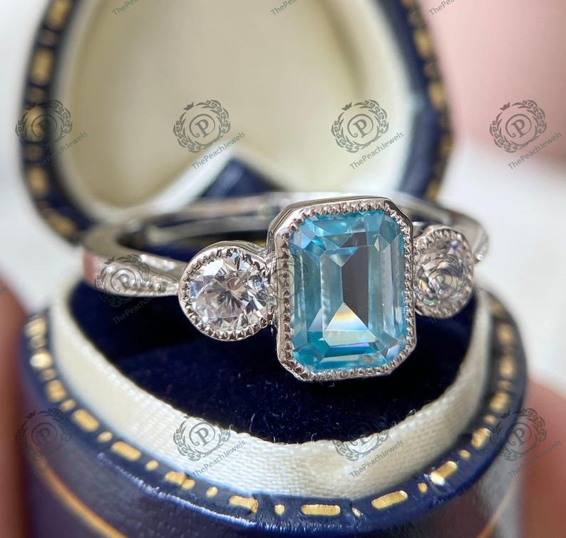 May include: A silver ring with a large, rectangular, light blue gemstone in the center, flanked by two smaller, round, clear gemstones. The ring is set in a blue jewelry box.