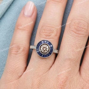 May include: A silver-toned ring featuring a central round champagne-colored stone surrounded by a halo of deep blue baguette-cut stones. The metal band has a polished finish and a circular setting that frames the center stone and blue accents.
