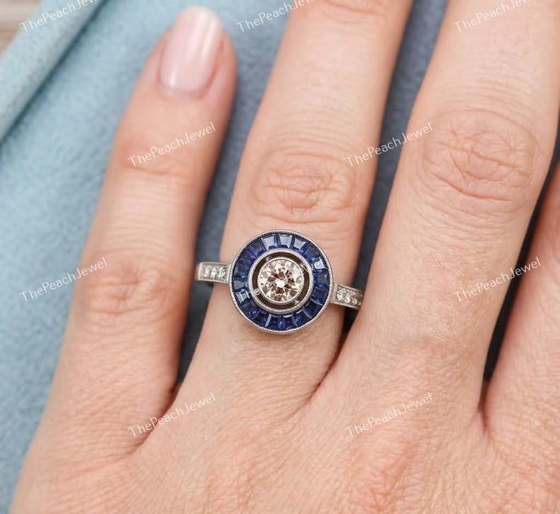 May include: A silver-toned ring featuring a central round champagne-colored stone surrounded by a halo of deep blue baguette-cut stones. The metal band has a polished finish and a circular setting that frames the center stone and blue accents.