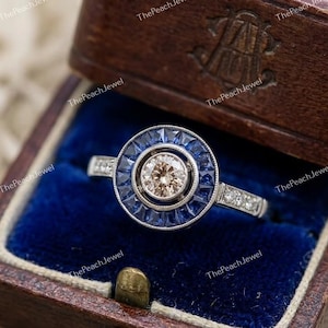 May include: A silver-toned ring featuring a central round champagne-colored stone surrounded by a halo of deep blue baguette-cut sapphires. The band is accented with small clear stones along the sides, set within a dark brown wooden jewelry box.
