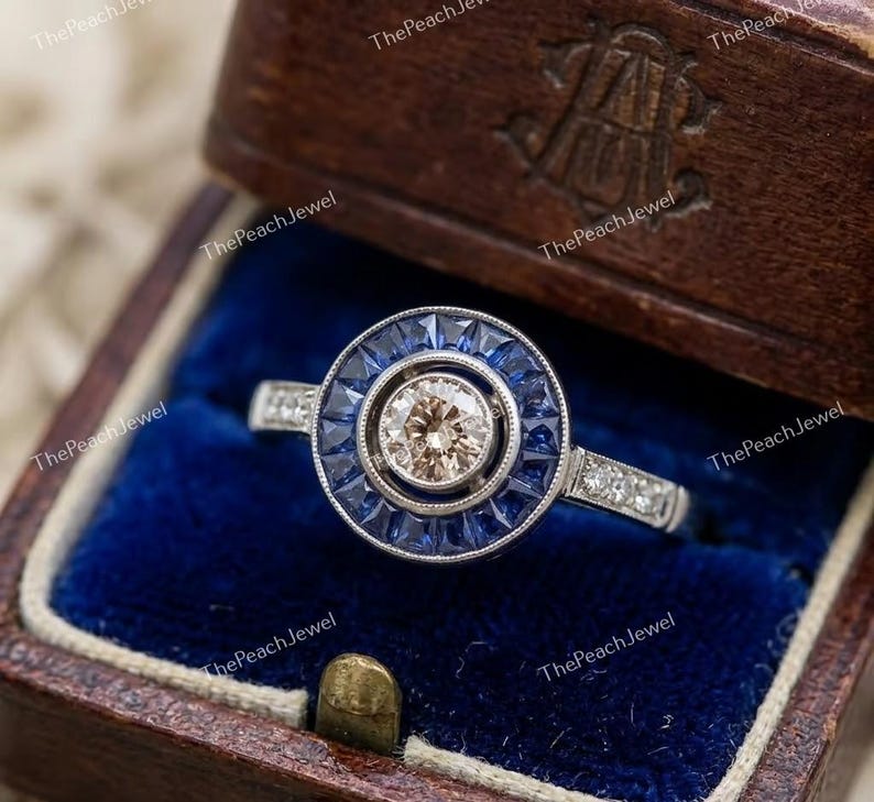 May include: A silver-toned ring featuring a central round champagne-colored stone surrounded by a halo of deep blue baguette-cut sapphires. The band is accented with small clear stones along the sides, set within a dark brown wooden jewelry box.
