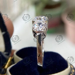 May include: A silver ring with three round, clear gemstones. The ring is set on a blue velvet background.
