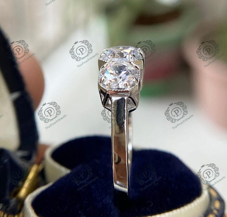 May include: A silver ring with three round, clear gemstones. The ring is set on a blue velvet background.