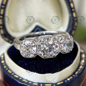 May include: A silver three-stone engagement ring with a large center diamond and two smaller diamonds on either side. The ring is set in a classic style with a milgrain finish.