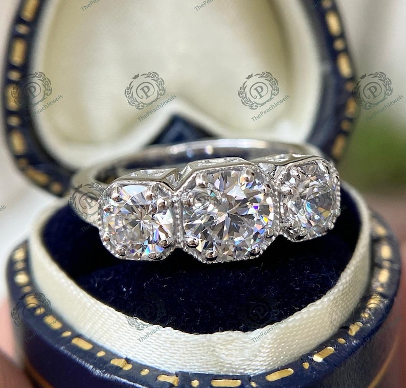 May include: A silver three-stone engagement ring with a large center diamond and two smaller diamonds on either side. The ring is set in a classic style with a milgrain finish.