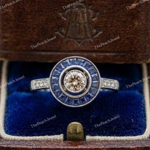 May include: A silver-toned ring featuring a central round champagne-colored gemstone surrounded by a halo of baguette-cut blue sapphires. The band is accented with small round clear stones on each side, presented in a dark brown box.