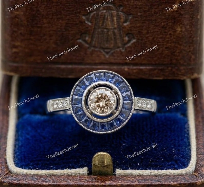 May include: A silver-toned ring featuring a central round champagne-colored gemstone surrounded by a halo of baguette-cut blue sapphires. The band is accented with small round clear stones on each side, presented in a dark brown box.