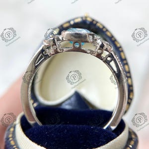 May include: A silver ring with a light blue gemstone, set in a decorative band. The ring is displayed in a blue velvet ring box. The ring has intricate detailing around the gemstone and along the band.