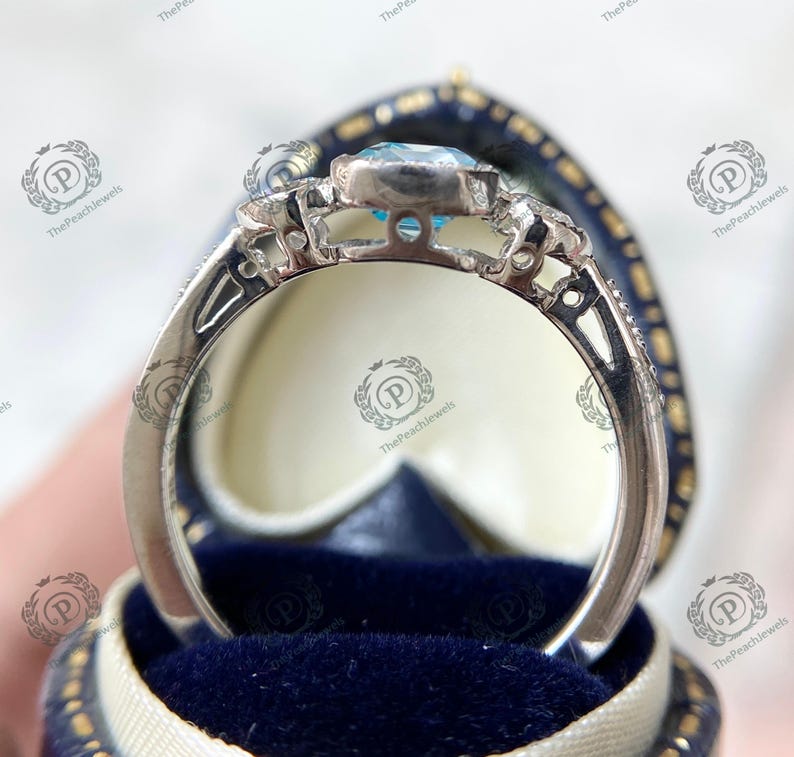 May include: A silver ring with a light blue gemstone, set in a decorative band. The ring is displayed in a blue velvet ring box. The ring has intricate detailing around the gemstone and along the band.