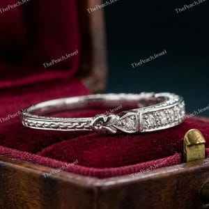 May include: A silver-toned metal band ring featuring an intricate engraved pattern along the shank and a series of small, clear, round-cut stones set into the front section, showcasing a detailed filigree-style design on the metal surface.