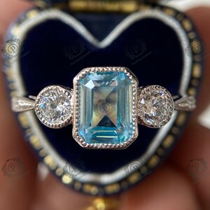 May include: A silver ring with a large, rectangular, light blue gemstone in the center, flanked by two smaller, round, clear gemstones. The ring is set against a dark blue, heart-shaped jewelry box.