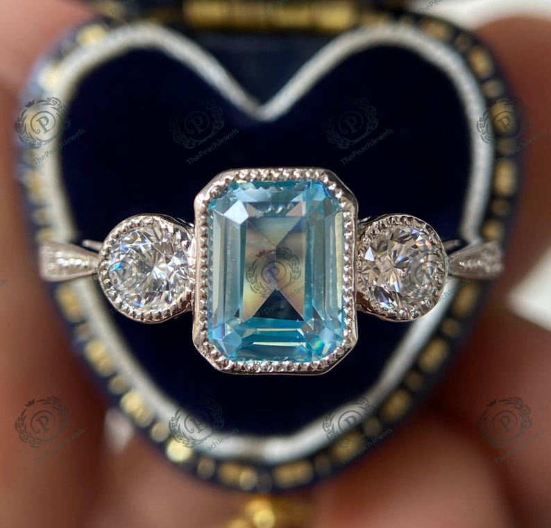 May include: A silver ring with a large, rectangular, light blue gemstone in the center, flanked by two smaller, round, clear gemstones. The ring is set against a dark blue, heart-shaped jewelry box.
