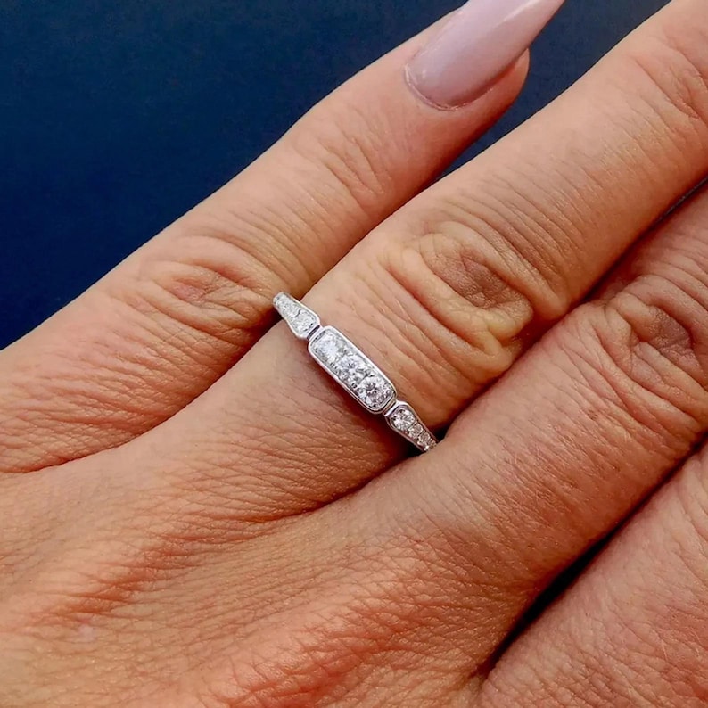 Puede incluir: Un delicado anillo de banda de plata con una fila de peque&ntilde;os diamantes transparentes engastados en forma rectangular. El anillo est&aacute; en un dedo.
