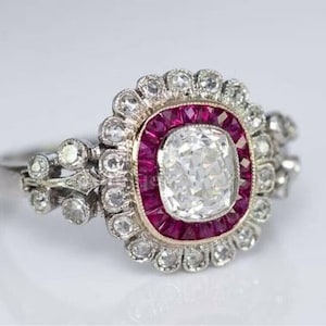 May include: A silver ring with a large, clear, cushion-cut diamond surrounded by a halo of smaller diamonds and a border of red rubies.