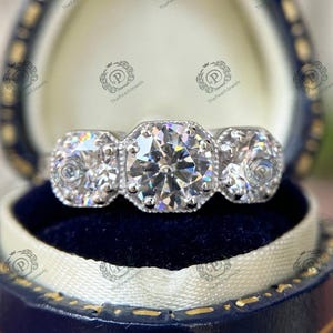 May include: A three-stone engagement ring with a large, round, center diamond and two smaller, round diamonds on either side. The ring is set in a white metal band.