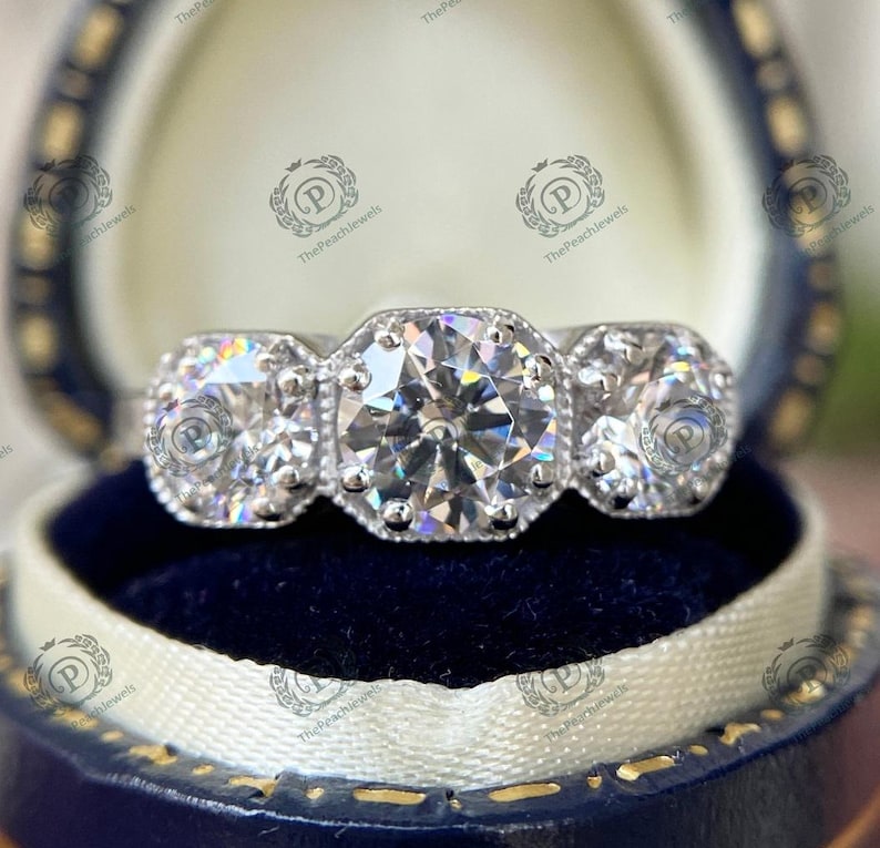 May include: A three-stone engagement ring with a large, round, center diamond and two smaller, round diamonds on either side. The ring is set in a white metal band.