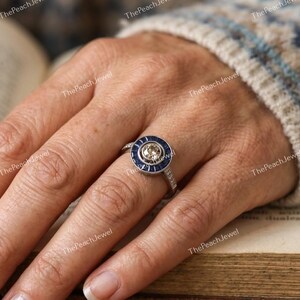 May include: A silver-toned ring featuring a central round champagne-colored stone surrounded by a halo of deep blue baguette-cut stones. The band is accented with small clear stones along the sides, creating a circular, multi-layered design.