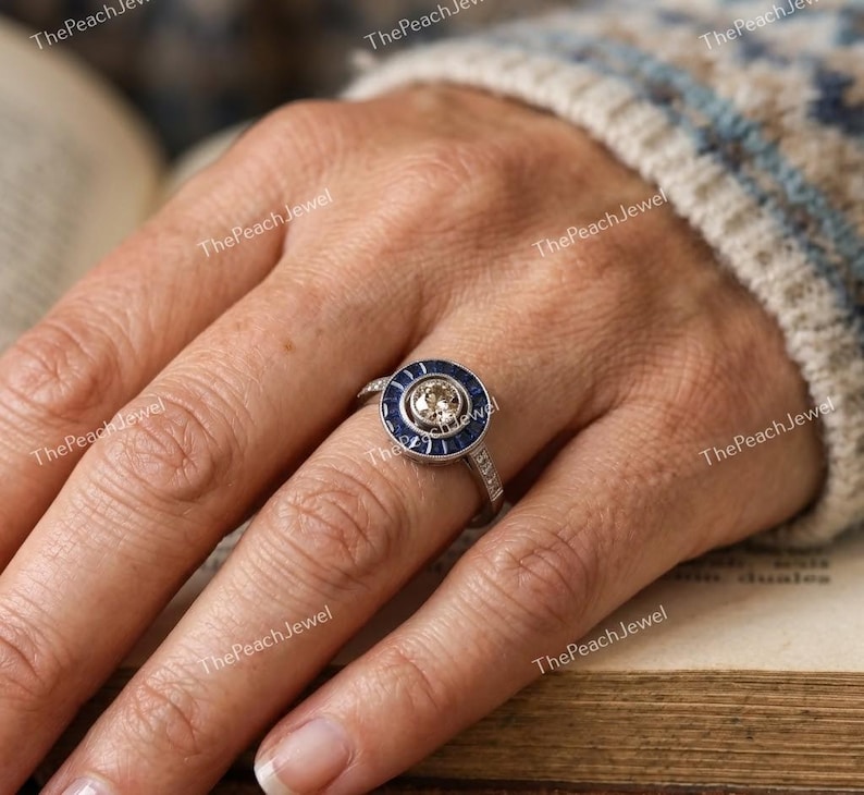 May include: A silver-toned ring featuring a central round champagne-colored stone surrounded by a halo of deep blue baguette-cut stones. The band is accented with small clear stones along the sides, creating a circular, multi-layered design.
