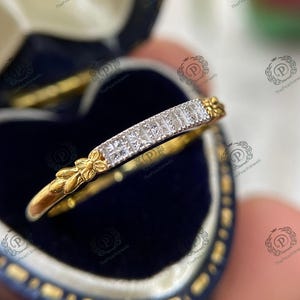 May include: A gold ring featuring a row of square-cut diamonds. The ring has floral and leaf detailing on the sides. The ring is presented in a blue jewelry box. This ring is a piece of jewelry.