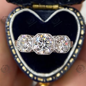 May include: A three-stone diamond engagement ring with a large center stone and two smaller stones on either side. The ring is set in a white metal band and is displayed in a blue velvet ring box.