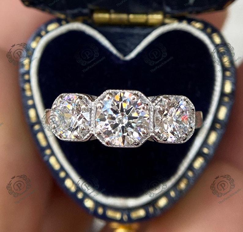 May include: A three-stone diamond engagement ring with a large center stone and two smaller stones on either side. The ring is set in a white metal band and is displayed in a blue velvet ring box.