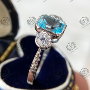May include: A silver ring with a large, rectangular, light blue gemstone and a smaller, round, clear gemstone. The ring is set in a decorative, ornate setting. The ring is displayed in a blue jewelry box.