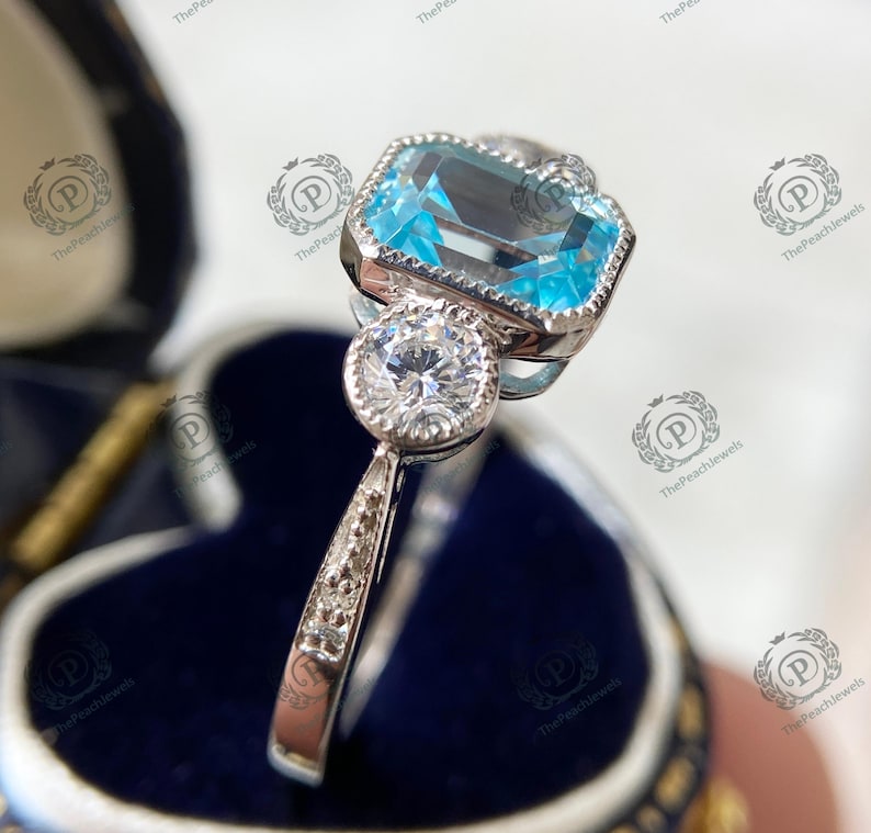 May include: A silver ring with a large, rectangular, light blue gemstone and a smaller, round, clear gemstone. The ring is set in a decorative, ornate setting. The ring is displayed in a blue jewelry box.