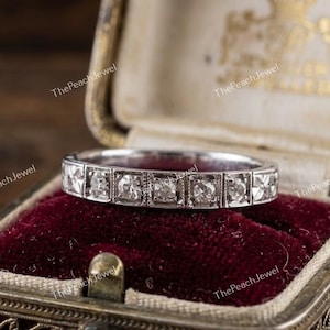 May include: A silver ring featuring square-cut diamonds in a channel setting. The ring is presented in an open vintage jewellery box with a red velvet lining. The box is ornate.