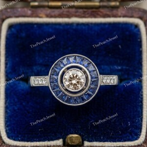May include: A silver-toned ring featuring a central round champagne-colored stone surrounded by a halo of baguette-cut blue sapphires. The band is accented with small round clear stones on each side, set in a circular frame with milgrain detailing.