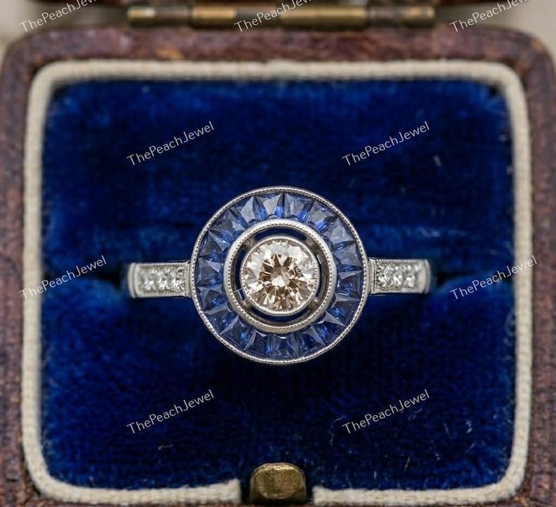 May include: A silver-toned ring featuring a central round champagne-colored stone surrounded by a halo of baguette-cut blue sapphires. The band is accented with small round clear stones on each side, set in a circular frame with milgrain detailing.