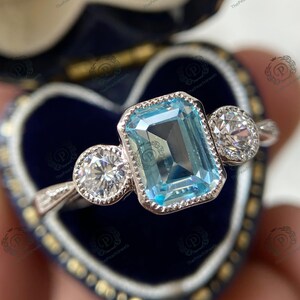 May include: A silver ring featuring a large, rectangular, light blue gemstone flanked by two smaller, round, clear gemstones. The ring is set in a blue velvet jewelry box.