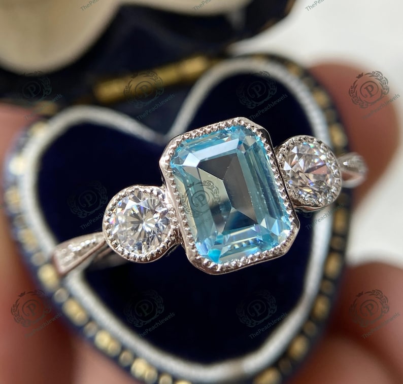 May include: A silver ring featuring a large, rectangular, light blue gemstone flanked by two smaller, round, clear gemstones. The ring is set in a blue velvet jewelry box.