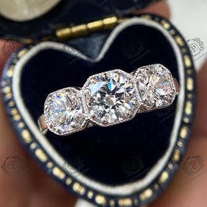 May include: A three-stone diamond engagement ring with a large center stone and two smaller stones on either side. The ring is set in white gold and has a classic, elegant design.