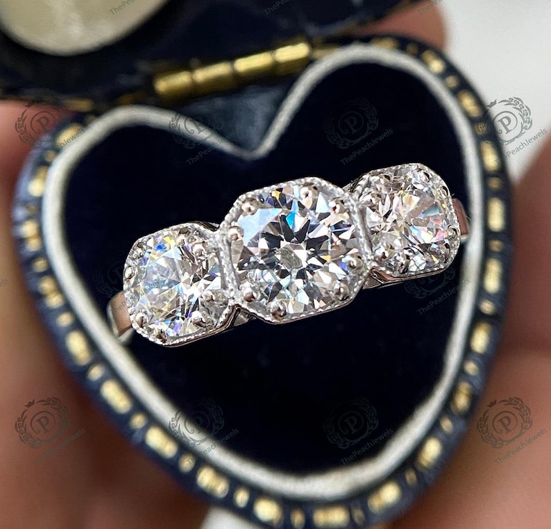 May include: A three-stone diamond engagement ring with a large center stone and two smaller stones on either side. The ring is set in white gold and has a classic, elegant design.