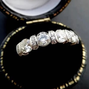 May include: A silver ring with five clear, sparkling gemstones set in a row. The ring is displayed inside a black jewelry box with a gold-colored trim, creating a striking contrast.