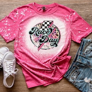 Puede incluir: Camiseta rosa blanqueada con un gráfico blanco de una bandera a cuadros y las palabras "Race Day" en una fuente retro.