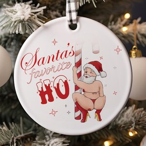 May include: White ceramic Christmas bauble featuring a red and white candy cane and a cartoon Santa Claus wearing a red hat and white beard. The bauble has the text "Santa's favourite HO" printed on it.