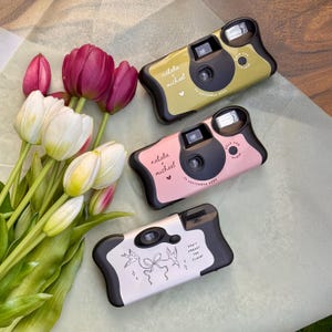 May include: Three disposable cameras with black accents and custom text. Two cameras are olive green and pink with the text 'natalie + michael' and '14 september 2025'. The third is white with a line drawing of two birds, a bow, and 'don't forget the flash'.