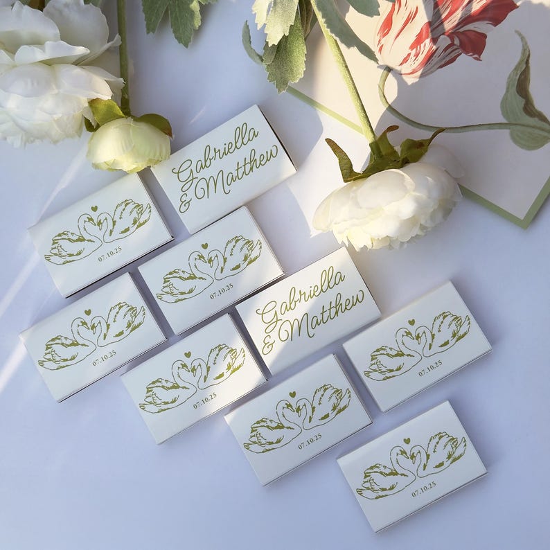 May include: A collection of rectangular white matchboxes featuring a gold foil print of two swans forming a heart shape, the date 07.10.25, and the names Gabriella & Matthew in a cursive script font, arranged on a flat surface with white flowers.