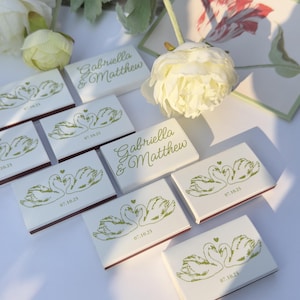 May include: A collection of rectangular matchboxes featuring a white surface with a green printed illustration of two swans forming a heart shape, the text Gabriella & Matthew, and the date 07.10.25, arranged alongside white floral accents.