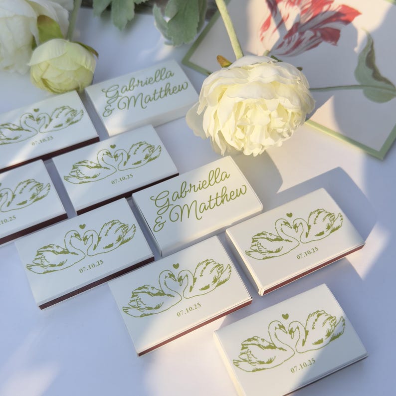 May include: A collection of rectangular matchboxes featuring a white surface with a green printed illustration of two swans forming a heart shape, the text Gabriella & Matthew, and the date 07.10.25, arranged alongside white floral accents.